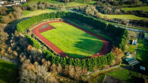 An aerial photo taken of the Whitchurch race track. There is a green field in the middle of the red oval running track and the track is surrounded by thick green trees and bushes.