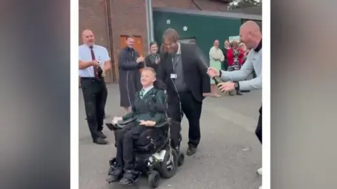 Patrick in his wheelchair and school uniform outside being applauded by a line of people.