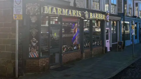 A new looking barbershop front with a gold lettering reading Marmaris Barber on a street with other shops continuing alongside. The are assorted traffic signs along the street and the barber shop has a traditional red, blue and white cylindrical spiral attached to its fascia.