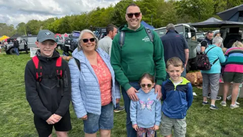 A teenager boy with a grey baseball hat and black top and short, left, a grey haired woman with a blue jacket, pink top and jean shorts, a man with a green hoody, sunglasses and a backpack puts his arms around his two small children, a boy and a girl. 