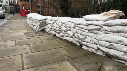 A low wall of white sandbags stretches for around 20 metres along the roadside, on Market Street. It is the height of seven sandbags. The Kyre Brook lies behind. 