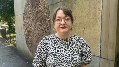 A woman with brown hair tied back stares at the camera. She wears a leopard print top, red lipstick, red glasses, gold earrings 