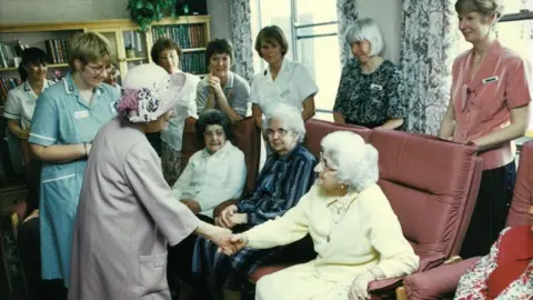 Cynthia Spencer Hospital The Queen Mother, shaking hands with a woman, who is sitting down on a red sofa. Are lot of people are in the room, looking at the woman and the Queen Mother. There are books on a shelf to the left. 