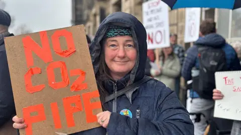 Protester Tina Lancaster wears a navy coat with the hood up and holds a sign which reads 'No Co2 Pipe' in orange writing outside Wallasey Town Hall. 