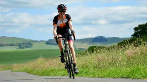 Supplied Alex Staniforth riding a bike. He is wearing a black helmet and outfit as well as sunglasses. He is smiling as he rides towards the camera. Green fields can be seen in the background.