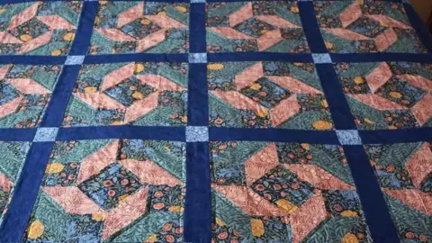Georgina Korn Quilt made up of square panels. Each panel is bordered by a thick dark blue strip. Where the strips meet there is a light blue flowery square. The panels themselves are covered in fabric featuring small flowers and leaves. These are arranged to form the shape of an angular flower.