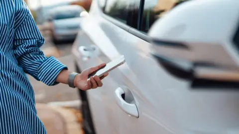 A woman is wearing a blue and white striped top and is holding a smartphone. She is standing next to a white car. 