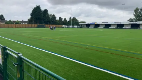 A wide view of the pitch and the stand.  The surface is bright green with newly painted lines.  There is a group of people standing on the pitch in the background.