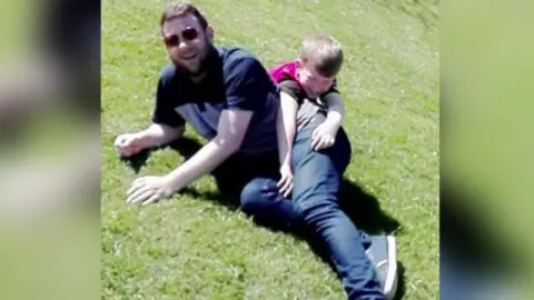 Alfie smiles as he rolls around with his father, wearing jeans, sunglasses and a black T-shirt, on a stretch of grass