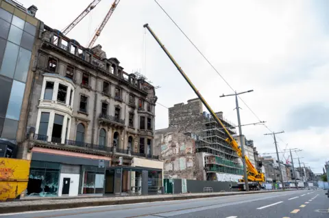 PA Media A general view of repair work being carried out on the former Debenhams building in Edinurgh. A large yellow crane is extended over the top of the building, which has been badly damaged and is charred black by a fire.