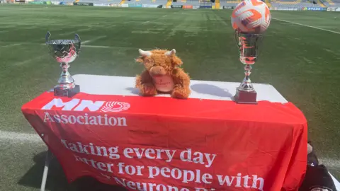 In the foreground, a table holds two trophies—one topped with a football—at either end, with a cuddly toy in the middle. A sports pitch is behind it and part of a stand is visible at the top of the photo.