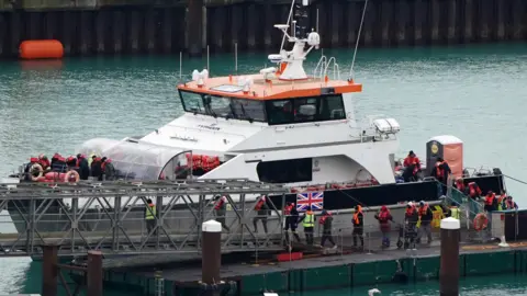 PA Media A white boat with an orange roof by the side of a jetty. Several people in dark clothes and lifejackets are disembarking.