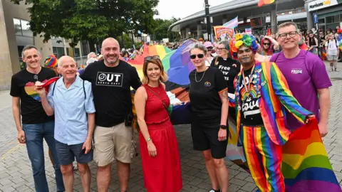 Haydn Brown Kim McGuinness is in the centre of the photo in a red top and matching skirt with a cross-body bag and sunglasses on top of her head. On the right is Kate Osborne who has grey, short hair and is wearing dark sunglasses, a black top and shorts. Next to her on the right is a man dressed in a rainbow suit with a rainbow flower hat and another man in a purple t-shirt and glasses. Lewis Atkinson stands on the far left and is wearing a black t-shirt with a rainbow heart on it and jeans. Between him and McGuinness are two men, one wearing a blue shirt and shorts with white hair, and a bald man wearing a black t-shirt which reads 'Out North East'
