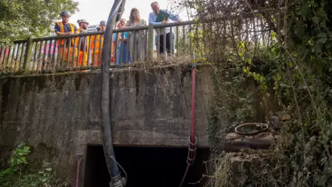 LDRS A group of people peer over a bridge. They are looking into a culvert. Men wearing high-vis gear and hard hats are with the group.