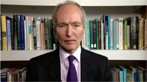 The photo shows a man dressed in formal attire, wearing a dark suit jacket, a light-colored dress shirt, and a purple tie. The background consists of a bookshelf filled with numerous books of varying sizes and colours. 