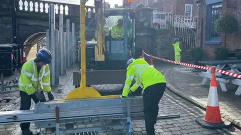 Environment Agency Two men in blue hard hats and high-vis jackets putting up flood defences, while another is in a mini digger lifting metal parts in to the pavement. An arch of a bridge is in the background