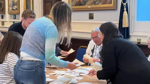 Several people sit and stand around a table while they count votes from the 2026 Jersey general election. Dozens of the ballots are on the table.