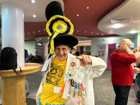 Nick the Incredible Flying Brick from the Monster Raving Loony Party, clad in a tall jaunty hat and lab coat with a rosette and other decorations