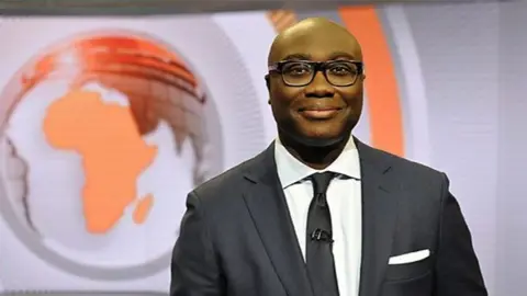 Komla Dumor, wearing a suit and glasses, smiles into the camera. BBC graphics can be seen on a large screen behind him.