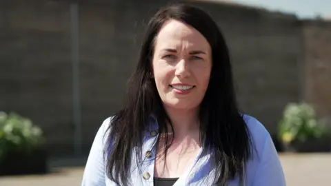 Lynsey Weston is standing outdoors and smiling at the camera. She has long, dark hair and is wearing a light blue buttoned top over a darker underlayer.