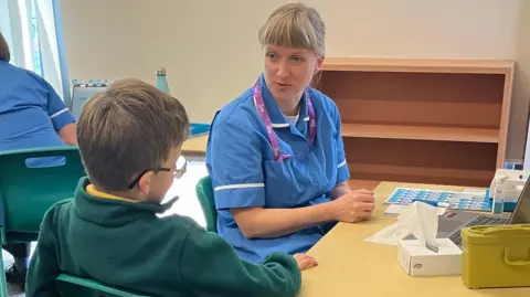 A nurse in a blue uniform sits at a desk talking to a boy of primary school age in a green sweatshirt and glasses. 