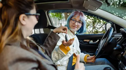 A stock image of a driving instructor sat in the passenger seat of a car, handing over the keys to the learner driver who is sat behind the wheel. The learner driver is wearing spectacles, is smiling, and has short blue and purple hair. She wears a white jacket over a yellow jumper, and a pair of jeans.