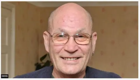 SWNS A man with short cropped hair and glasses smiling. He is wearing a blue top. In his left ear is an earring.