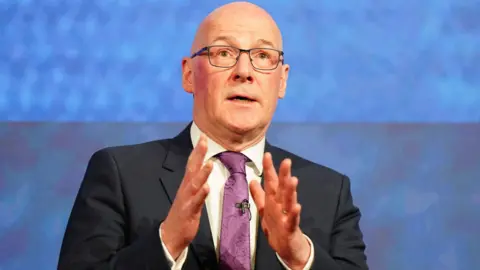 PA Media John Swinney wearing a dark suit over a white shirt and purple tie. He is standing in front of a blue background. His hands are raised in front of his chest.