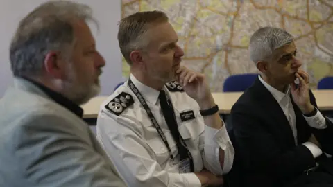 Matt Jukes and the Mayor sit beside watch other, listening with their hands on their chins, with a map of London in the background