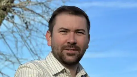 Callum Ringer has short brown hair and beard and is standing in the middle. He is wearing a white and black chequered shirt and is standing in front of a tree.