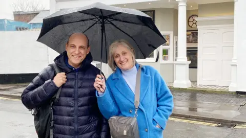 Christopher Baines Christopher Baines and Helen Sheppard smiling for the camera outside, while it's raining. She is holding a black umbrella. They are both wearing thick coats. The street behind them is wet.