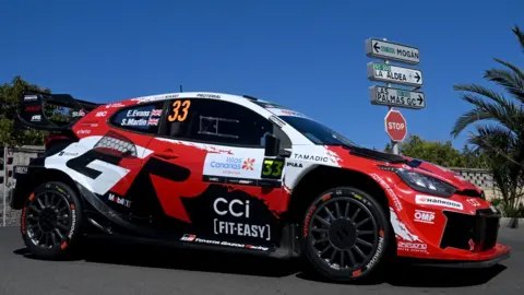 Elfyn Evans' red and white Toyota Yaris with blue sky, road signs and palm trees at Rally Islas Canarias