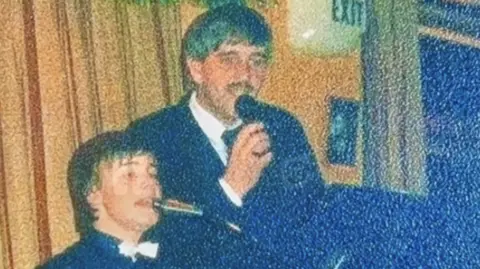 Alan Davies A young Gary Barlow on stage in front of a piano with Alan's dad stood by him and the piano. They are both singing into microphones and wearing shirts and blazers.