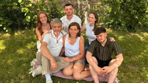 Family Handout The man and woman from the first photo, sitting on a blanket on the grass. It is sunny but they are sitting in shade. They are surrounded by their four children, two girls and two boys, all of whom appear to be in their late teens or early twenties.