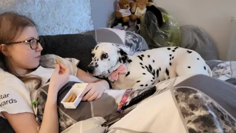 Publicaciones de Cancer Research UK Sophie Claxton, de pelo largo y castaño, yace en el sofá, debajo de un edredón, comiendo palitos de pan y un refrigerio de queso tierno mientras su dálmata de manchas blancas y negras, Bonnie, se acuesta a su lado y mira fijamente el refrigerio. Bonnie lleva una pajarita rosa, blanca y negra.