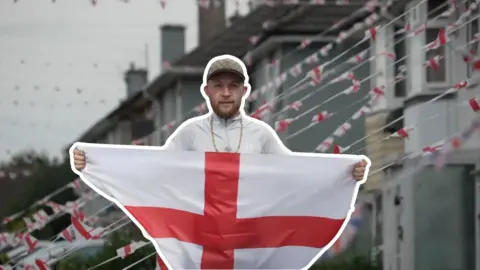 A man stands with an England flag outstretched in his arms. He has a beard, cap and gold chain around his neck. Behind him is a blurred picture of a row of houses with a string of England bunting draped from them. 