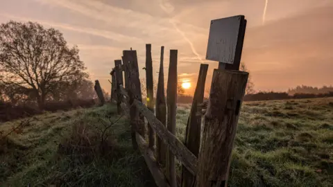 BBC Weather Watchers/alideakin A rough-cut wooden fence with a weather-worn sign on it is in the foreground of a shot with a dewy or frosty field and the sun rising over the horizon.