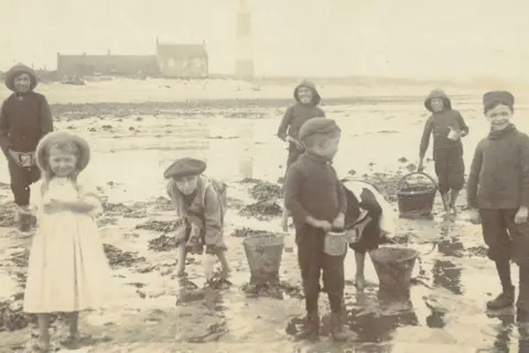 East Riding Archives and Local Studies A sepia-tinged black and white photo of young children standing on a beach with buckets, dressed in the clothing of the late Victorian era. Several boys are wearing hats and collecting stuff in the buckets and a girl in a white dress stands to the side, with a woman stood behind her. In the distance you can see a house with a lighthouse next to it.