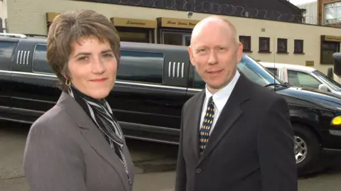 Julie and Rod stand in front of a stretch limo and they are both dressed in a chauffeurs' outfit of a dark jacket, shirt and Rod has a tie on. Julie has short dark hair and Rod has short ginger hair. They are both smiling at the camera.