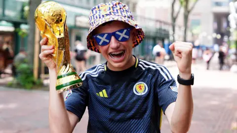 PA Media Scotland fan Richard Harvey holds a replica World Cup Trophy in Boston, Massachusetts. He is wearing a blue Scotland top, sunglasses with a saltire pattern, and a blue, pink and yellow patterned bucket hat.
