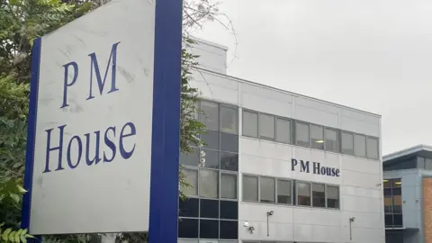 A large office building with the name “PM House” displayed prominently on its exterior. The building has a modern, rectangular design with multiple rows of windows and light grey cladding. In the foreground, a tall sign also reads “PM House” in blue lettering on a white background, framed by a dark blue border. Several cars are parked in front of the building, suggesting a business or office complex. 