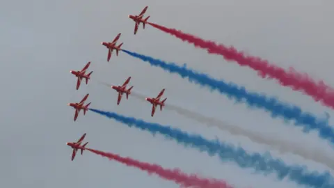 The red arrows in the sky at the Guernsey Air Display. There are seven planes with red, blue and white smoke coming from them. 