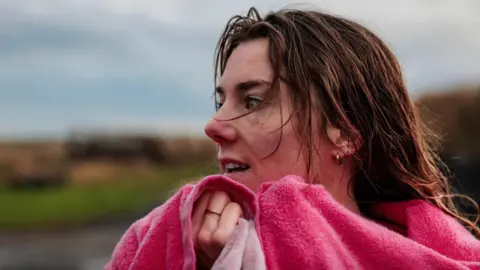The image shows a close‑up outdoor portrait near the coast of a person with wet hair, wrapped in a bright pink towel and holding it up around their neck. The background is softly blurred, showing a muted coastal landscape with grass, dunes and an overcast sky.