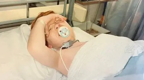 Kieran Cooper A child lies in a hospital bed. He is holding his hand to his forehead. He has a dummy in his mouth.