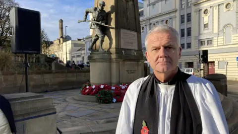 A clergyman wearing a white cassock and a black stole, with a poppy pinned on it. He is standing in front of a Cenotaph with poppy wreaths laid at the foot of the monument.