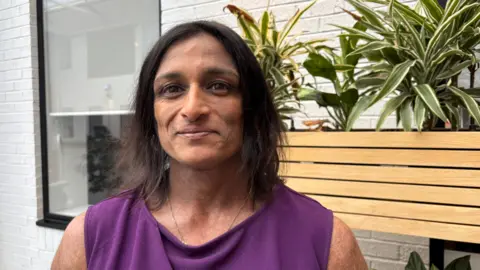 Martin Heath/BBC Ajanta Hilton with long dark hair smiling at the camera while wearing a purple dress. She is standing in front of a wooden planter with long-leaf green plants set in it. There is a window with a half-closed blind to her right.