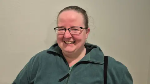 Chery Rowland smiles into the camera. She has glasses, short, dark hair and a green jacket on.