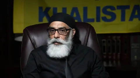 Bloomberg via Getty Images man with long white beard and glasses, wearing a tie and button-down in front of a flag that says "Khalistan"