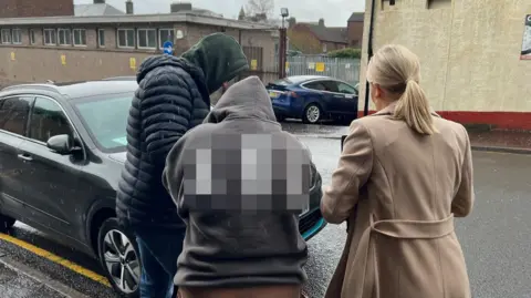 A woman with a hooded top with her back to the camera is taken to an unmarked police car by two plain clothes officers