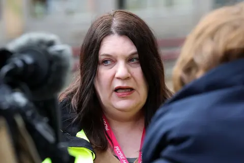 Getty Images Susan Aitken, a woman with long dark hair, is standing among media, being interviewed outside.